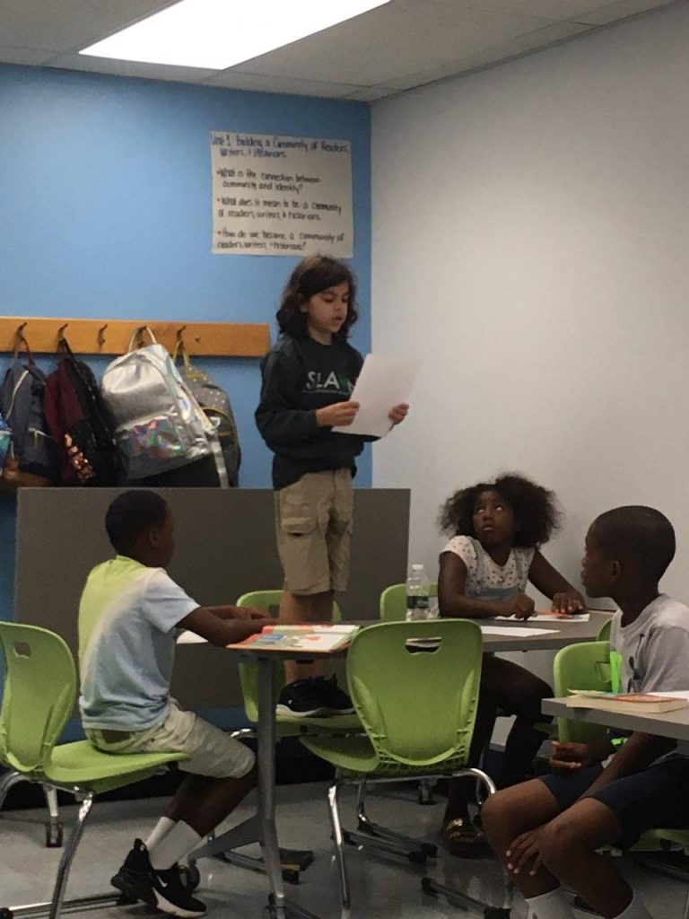 Student standing on chair reading aloud to classmates